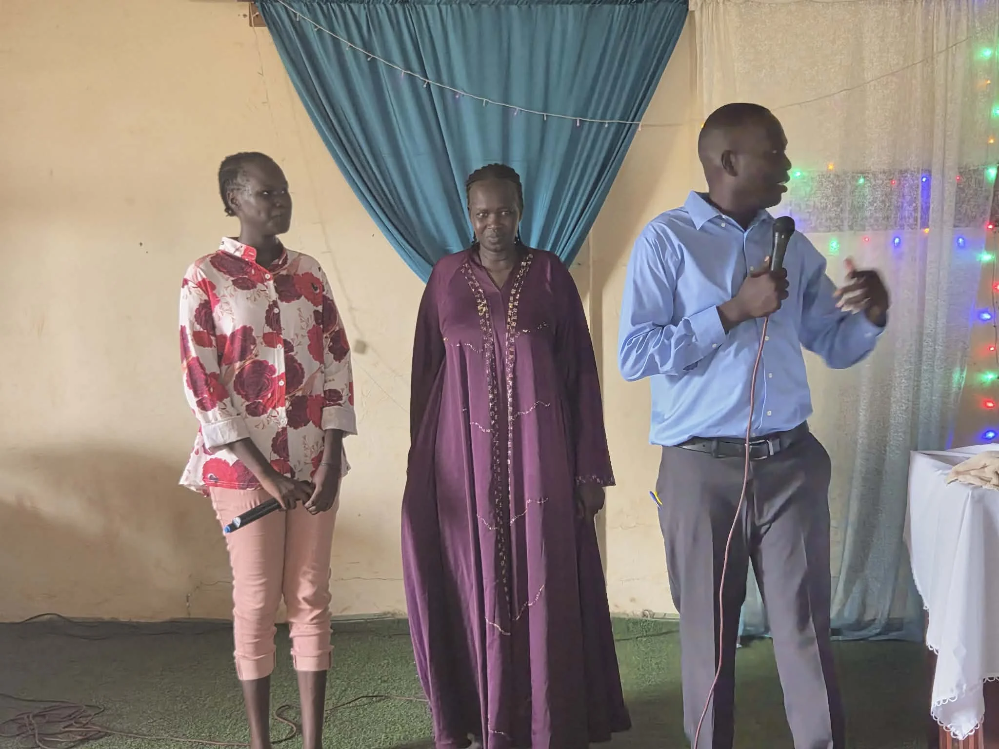 Three women and one man standing indoors against a yellow wall with blue curtain and decorative lights. The woman on the left is wearing a floral shirt, the woman in middle is dressed in purple traditional attire, the woman on the right is wearing gl