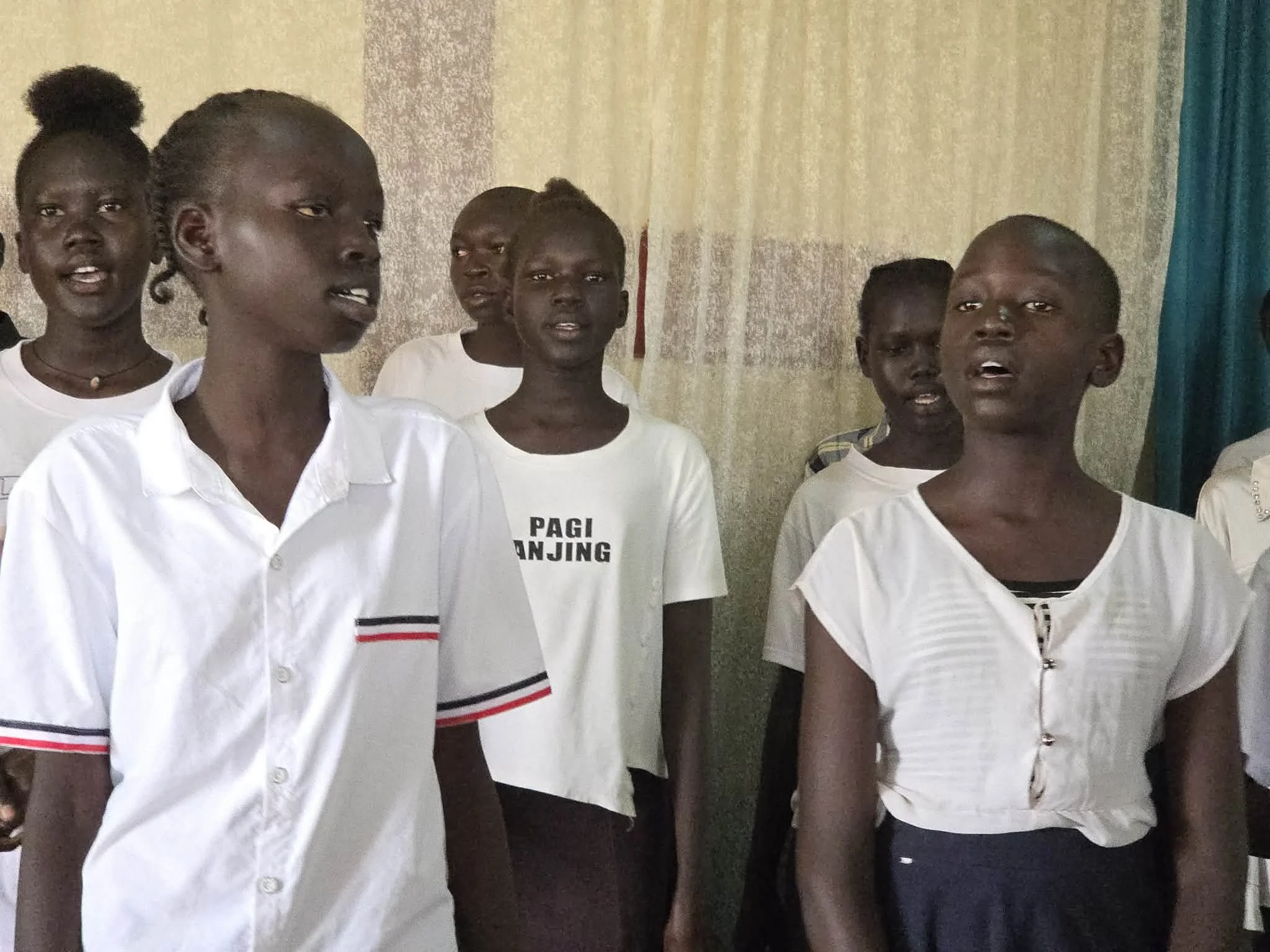 Group of children standing indoors, some wearing white shirts, in a room with curtains.