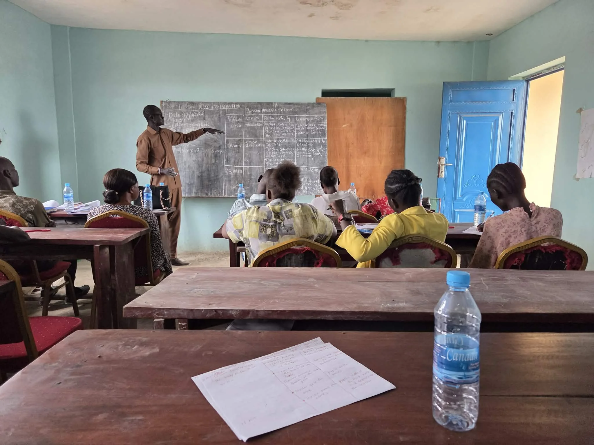 A classroom with students seated at tables facing a chalkboard. A teacher is standing next to the chalkboard, pointing at it. The room has light green walls and a blue door. Bottled water and papers are on the tables.