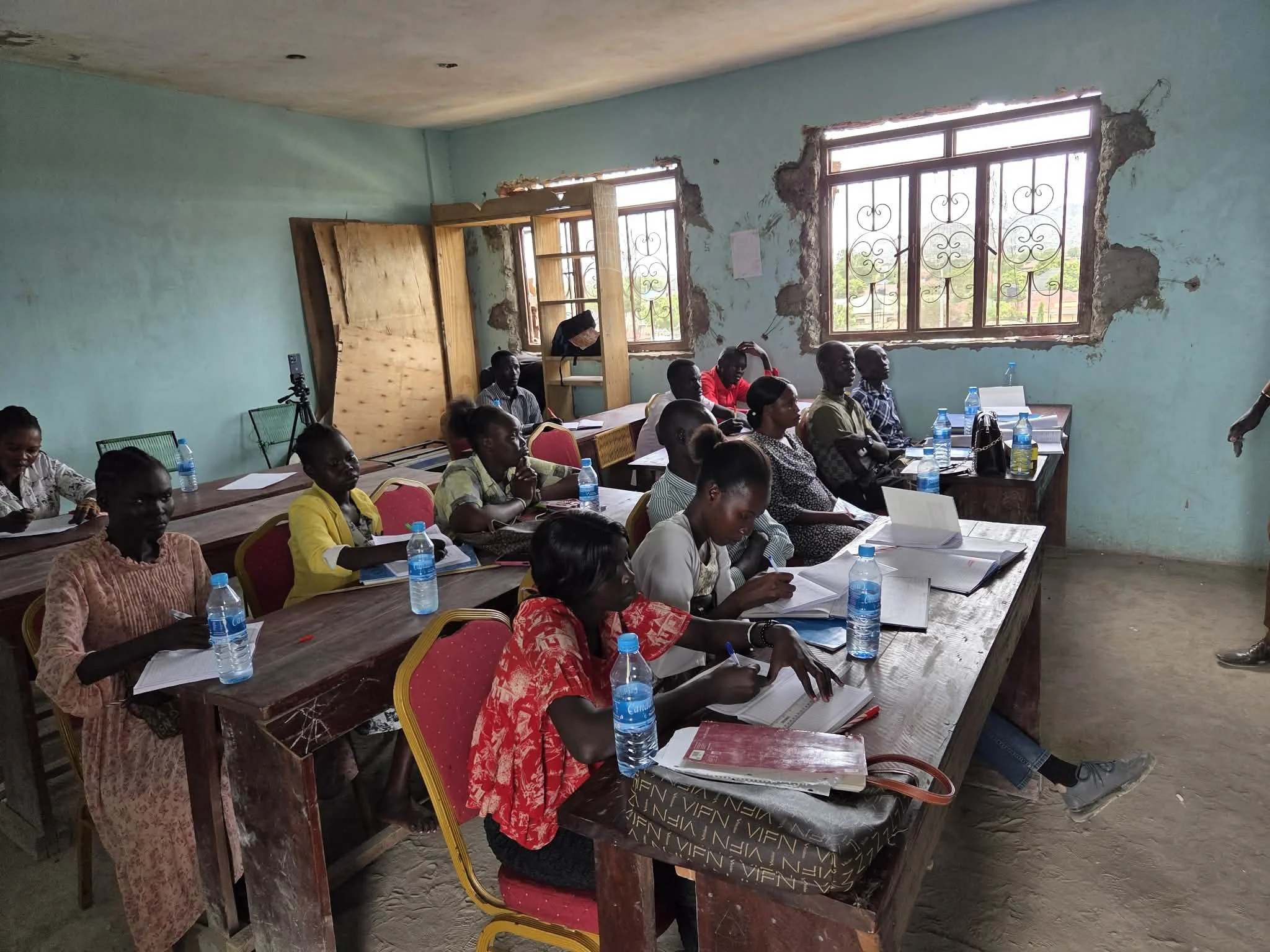 A group of men and women seated in a classroom or meeting room with desks, notebooks, and bottled water, listening attentively. The room has two large windows with decorative metal bars, and the wall behind them is damaged with exposed concrete. The 