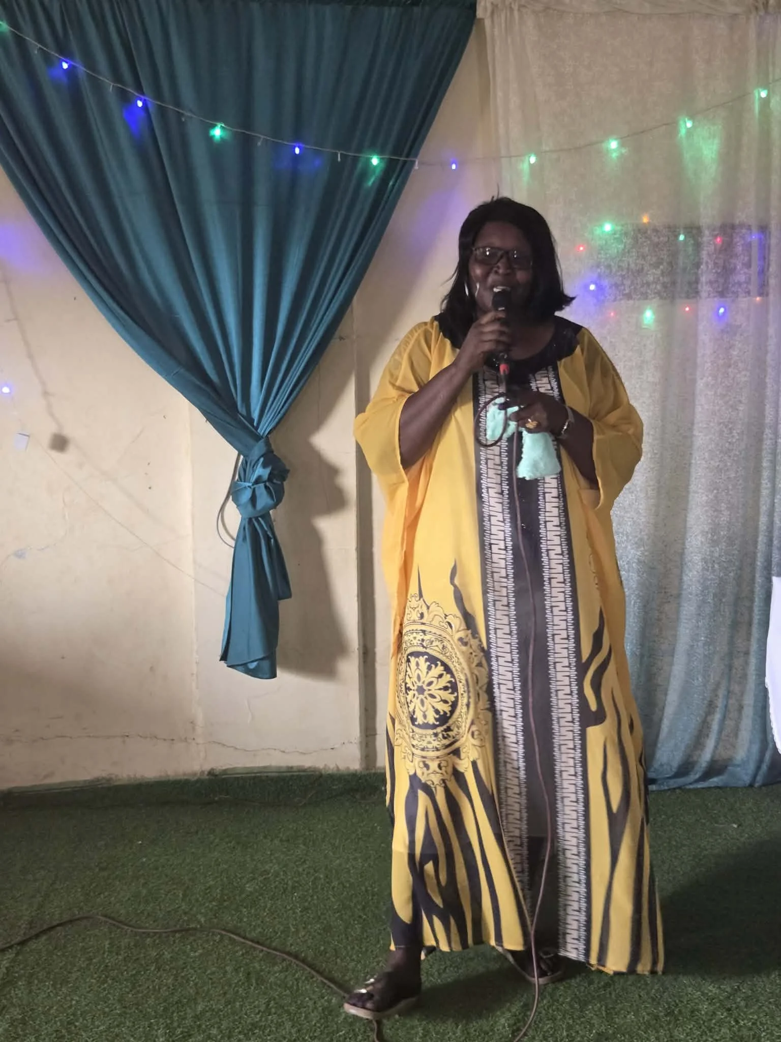 Woman in a yellow and black traditional dress, holding a microphone, standing on green carpet with a backdrop of colorful lights.