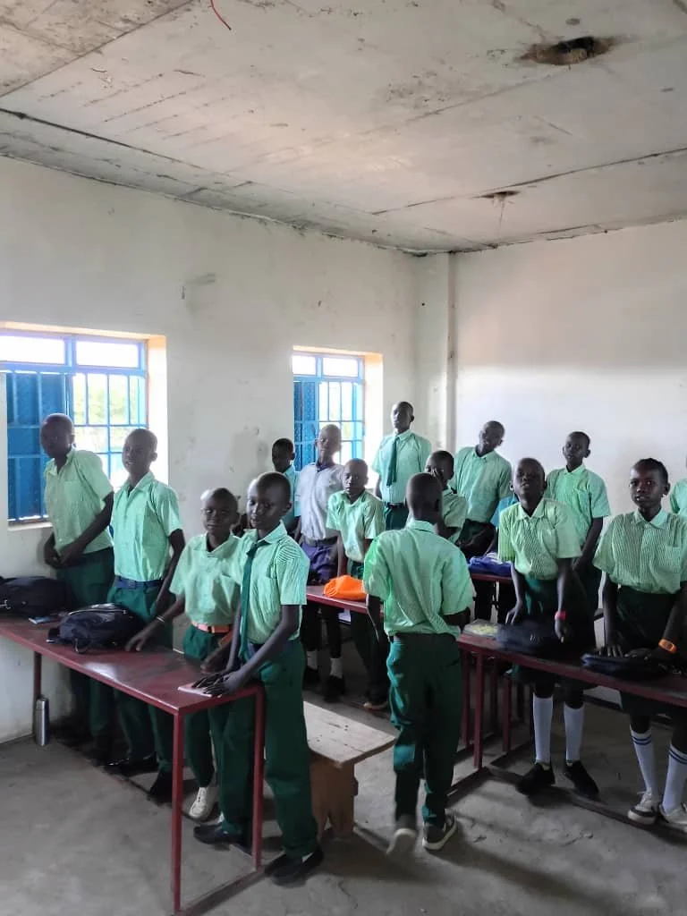 Students in green uniforms standing in a classroom with blue window bars and white walls.