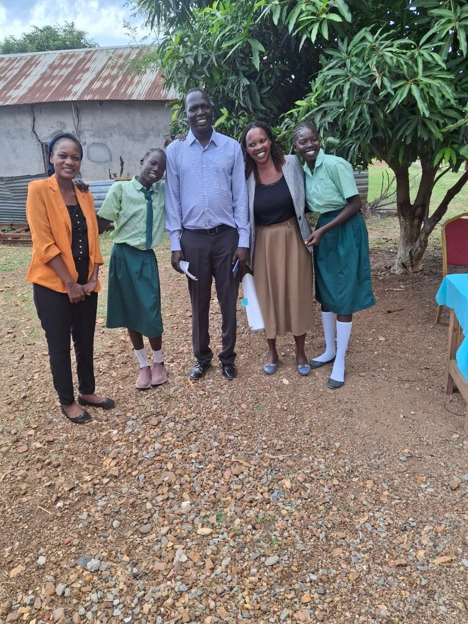 Five people, including two women and three children, standing outdoors on a dirt ground near a large leafy tree. The background shows a small building with a rusty roof. All are smiling and dressed semi-formally.