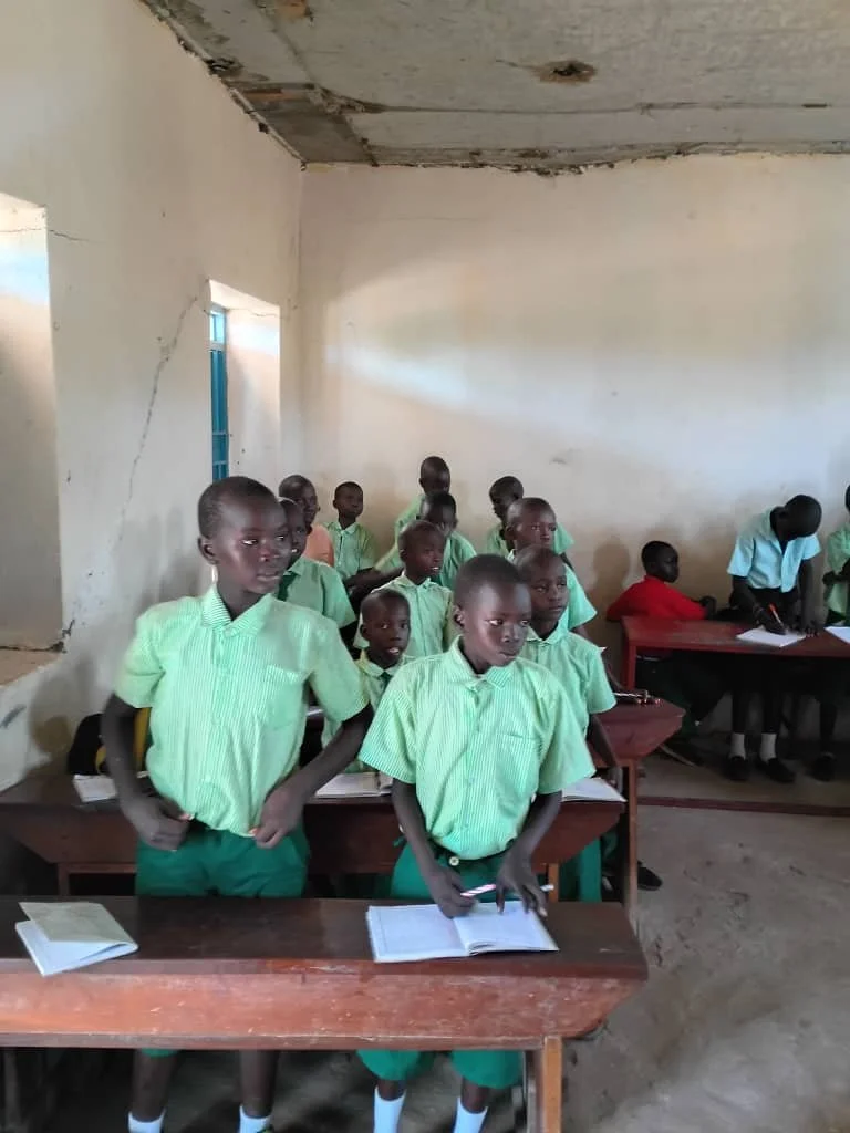 School classroom with young students in green uniforms, some standing and some seated at desks, with open books, inside a simple room with plain walls and a damaged ceiling.