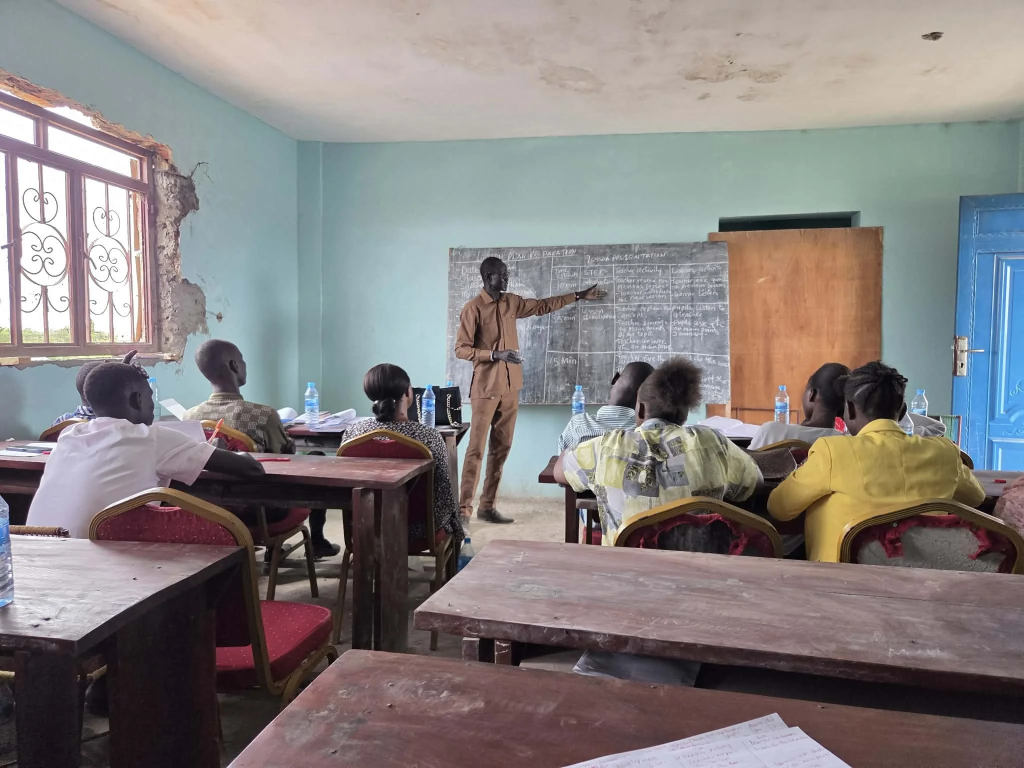 A classroom with students seated at wooden desks, facing a chalkboard where a teacher is pointing; a window with bars and a partially damaged wall frame is visible in the background.