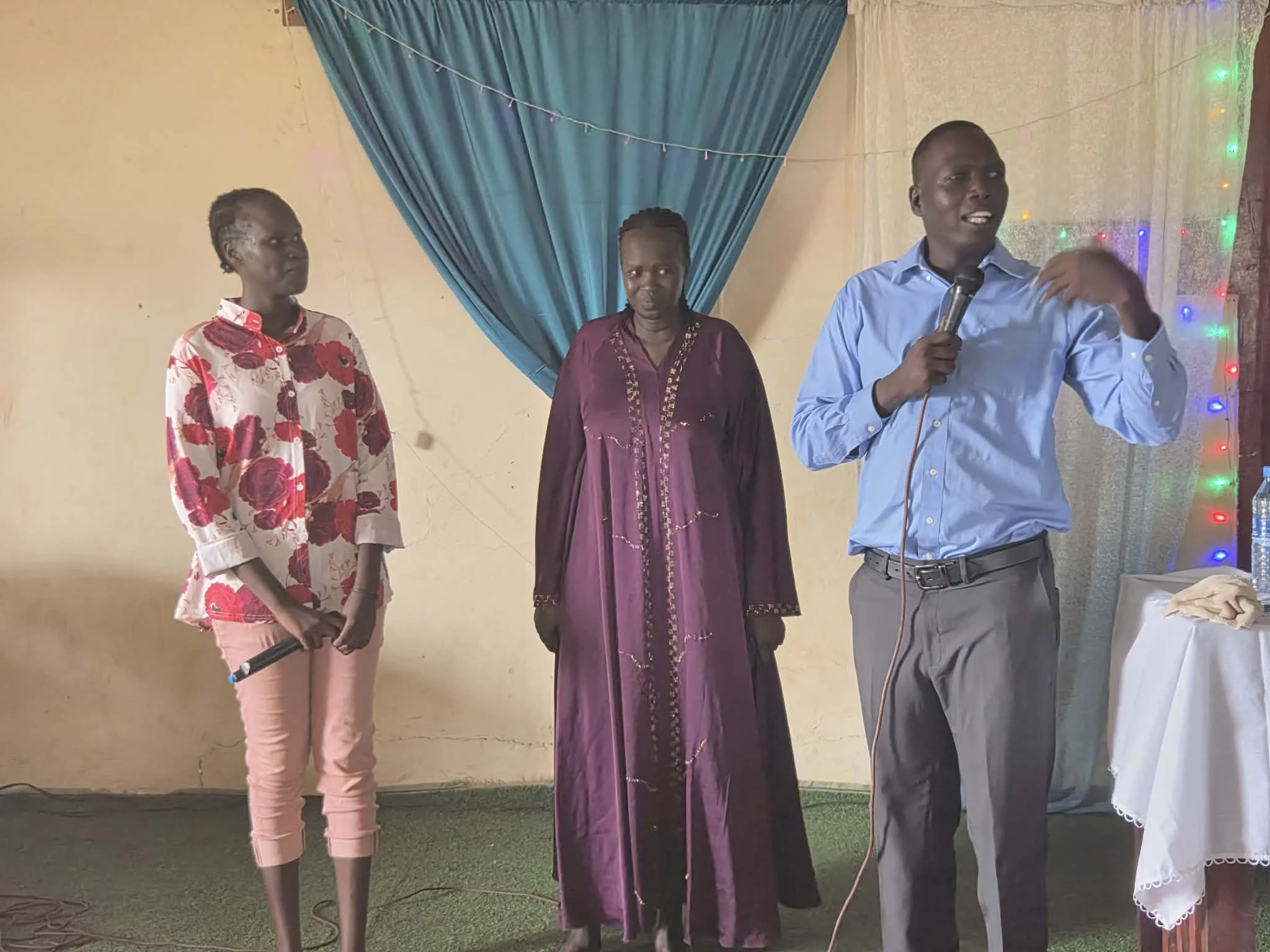 Four people standing indoors, with one person speaking into a microphone. The background includes blue curtains and colorful string lights, with a beige wall.