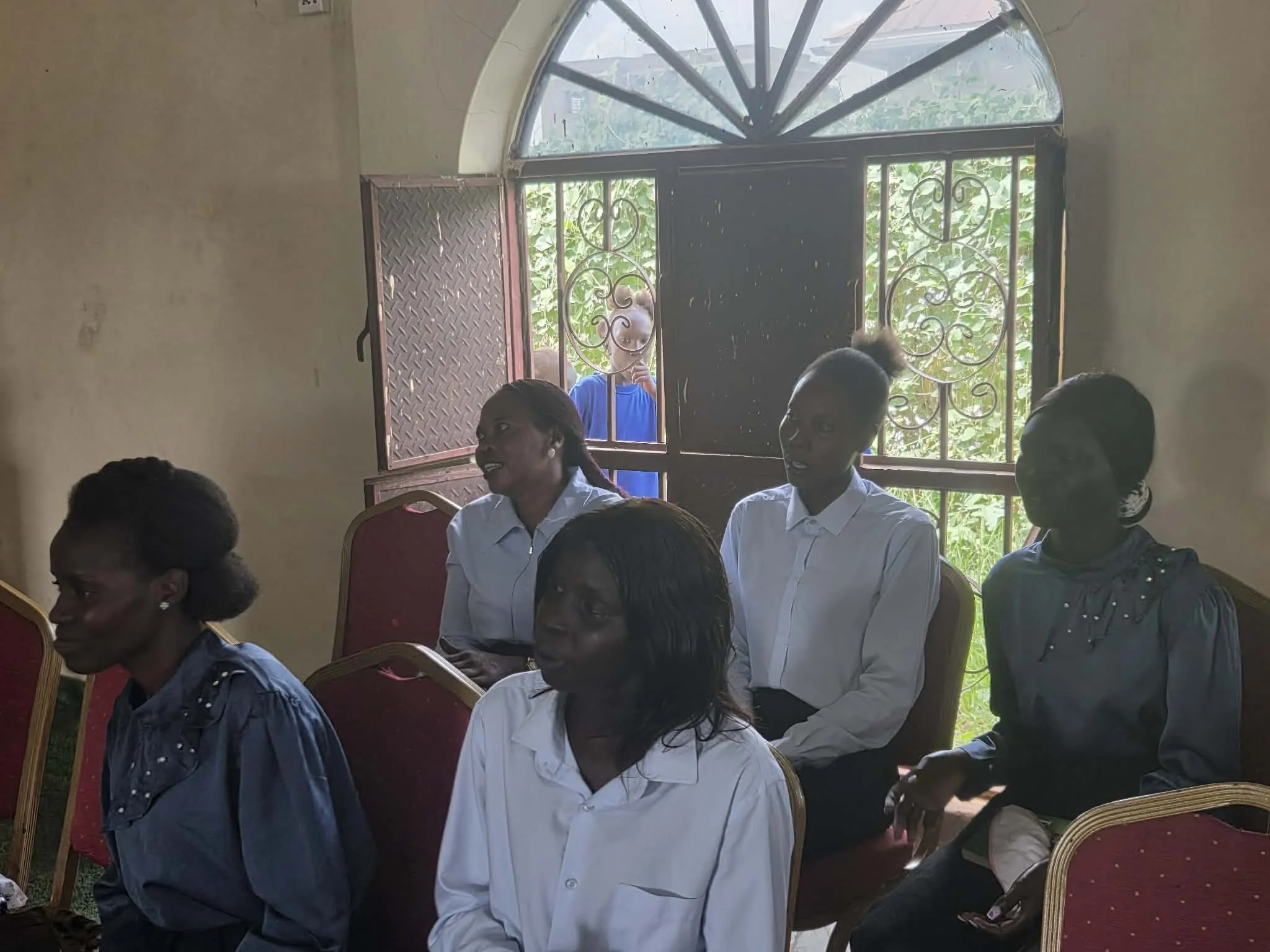 People sitting indoors near open window with decorative iron grilles, some women in white shirts, one woman in blue, a person standing outside the window.