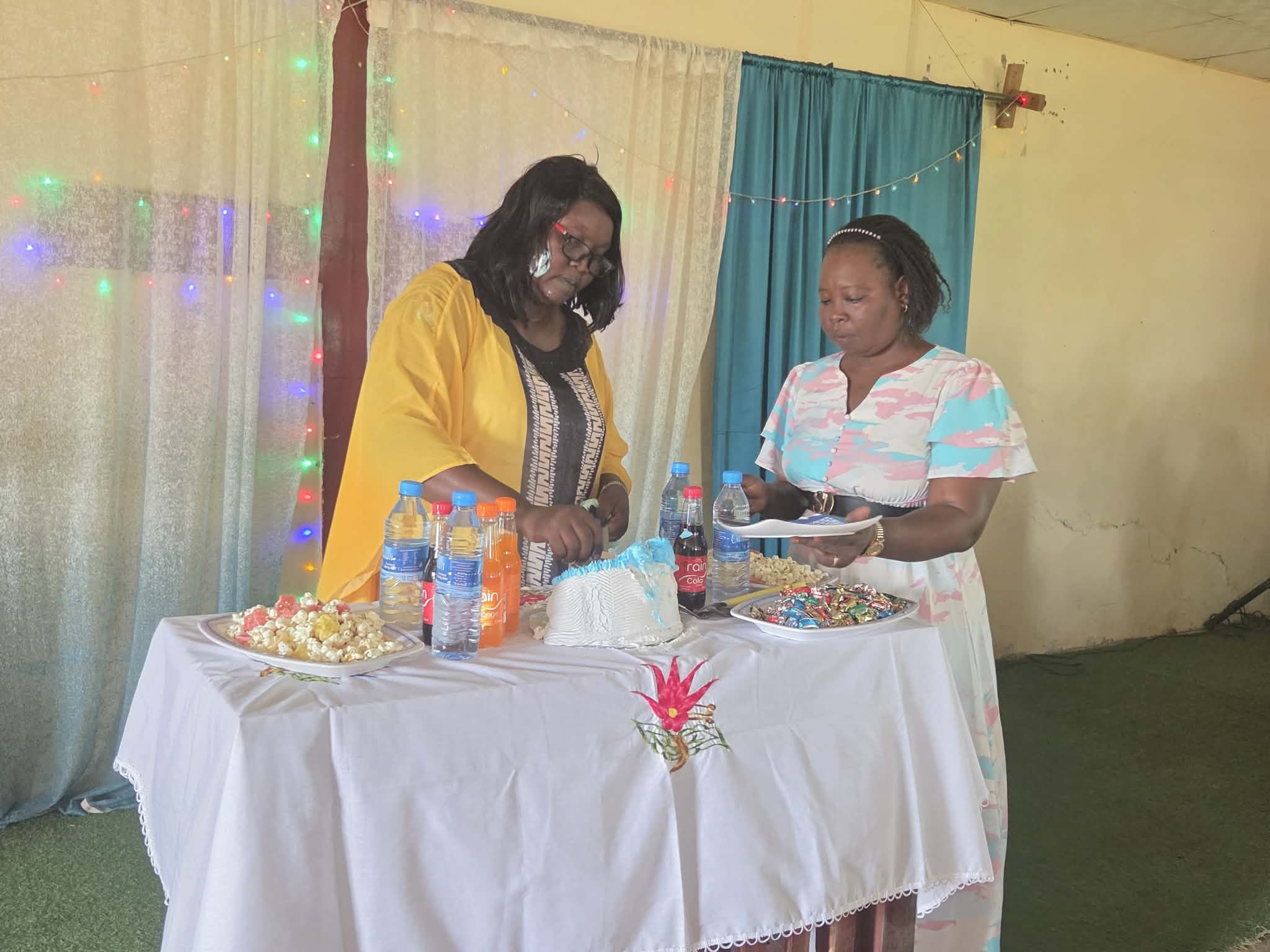 Two women standing at a decorated table with snacks, drinks, and flowers, engaged in a moment of prayer or reflection indoors with colorful string lights and curtains in the background.