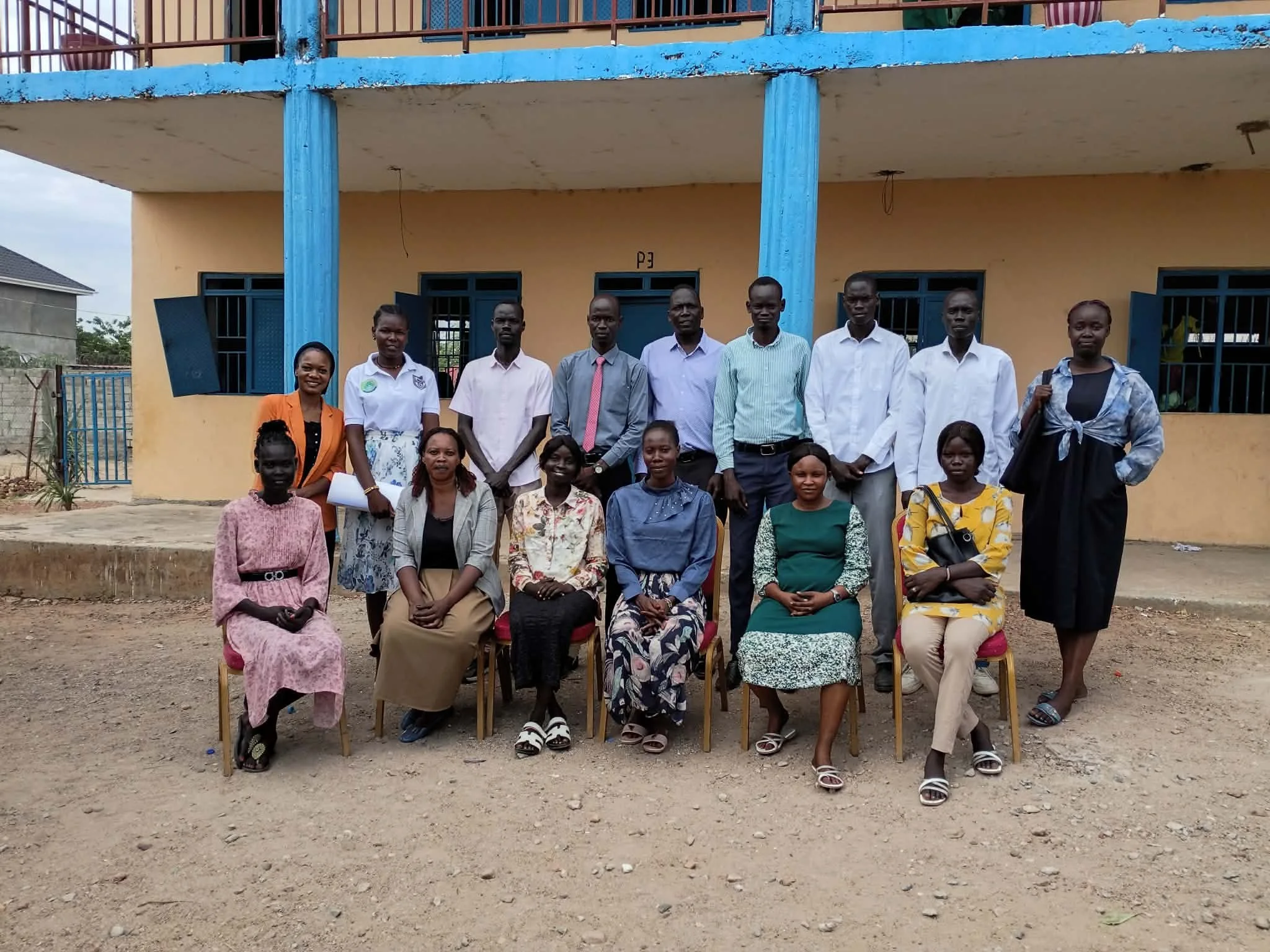 A group of men and women, some standing and some sitting, posing for a photo in front of a school building with blue columns and yellow walls. The people appear to be of African descent.