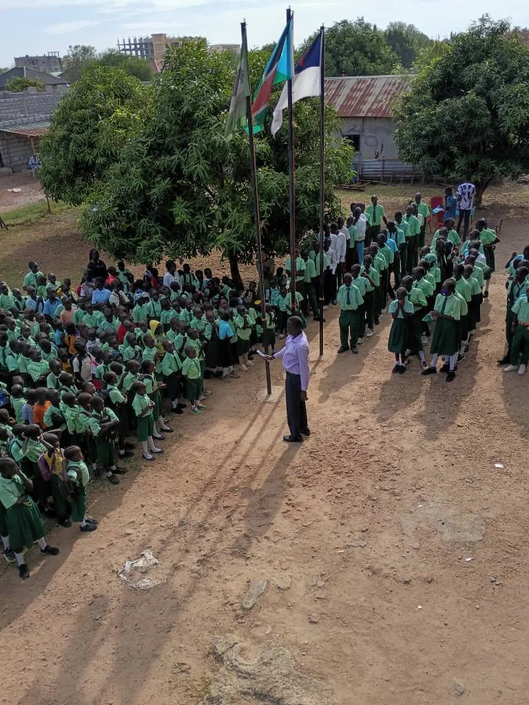 School assembly with students in green uniforms standing in lines under a tree, with a teacher or school official in a white shirt holding a document, near flagpoles with flags. The area is a dirt yard with some buildings and trees in the background.
