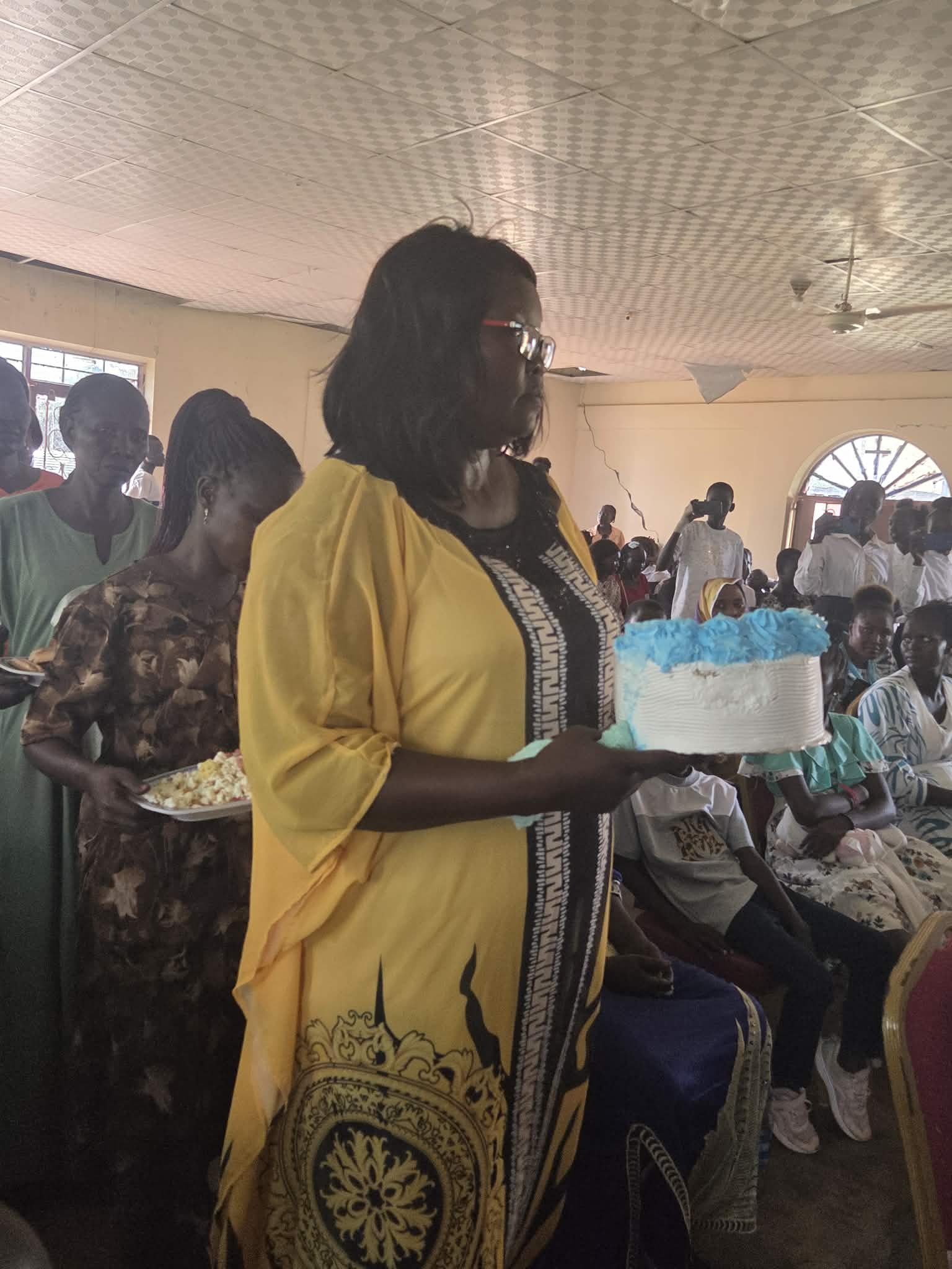 A woman in a yellow and black traditional outfit holding a cake with blue decorations at a gathering of people in a room with a patterned ceiling and large window.