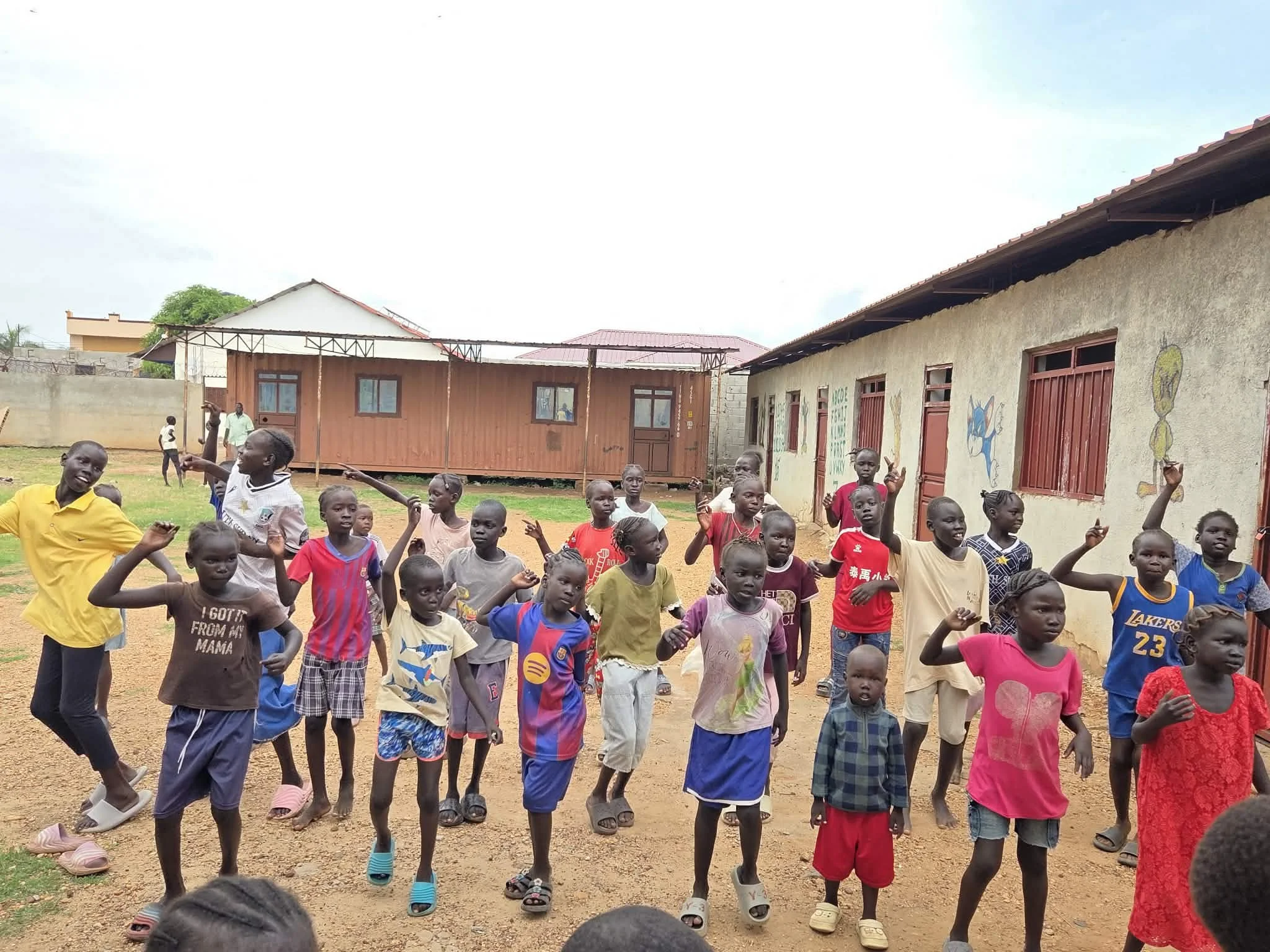 Children playing outside a school building with colorful murals, in a rural setting with dirt ground and simple structures.