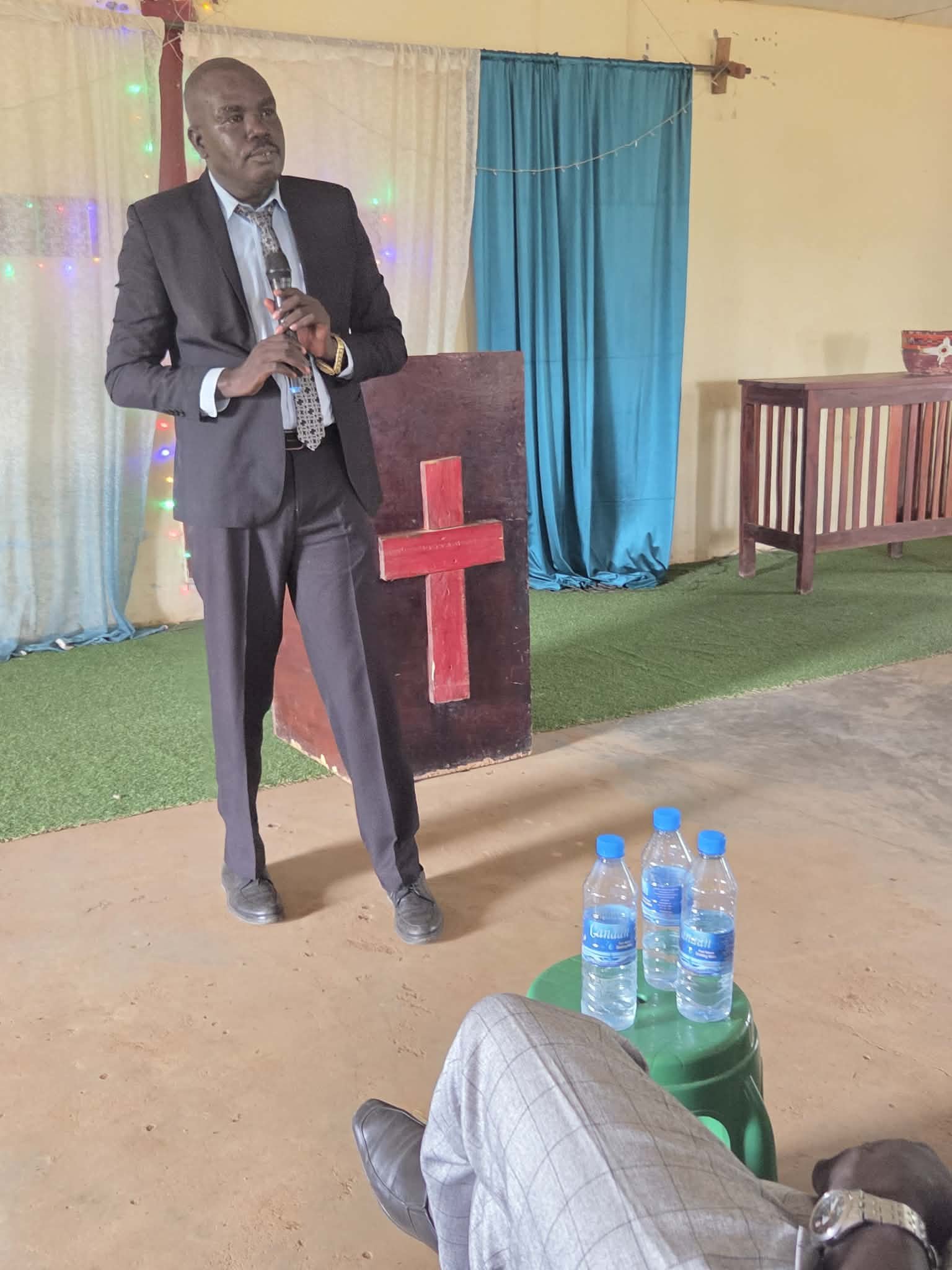 A man in a suit holding a microphone, standing in front of a makeshift altar with a red cross on a black panel, in a room decorated with blue curtains and multicolored lights. Three bottles of water are on a small green table in the foreground.