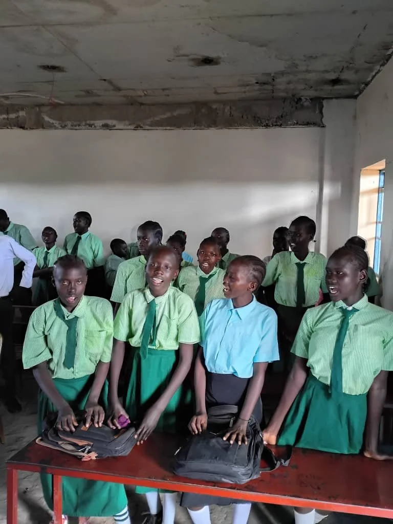 Group of school children in green uniforms with two girls in front, one in a blue shirt, in a classroom with a partially finished ceiling.