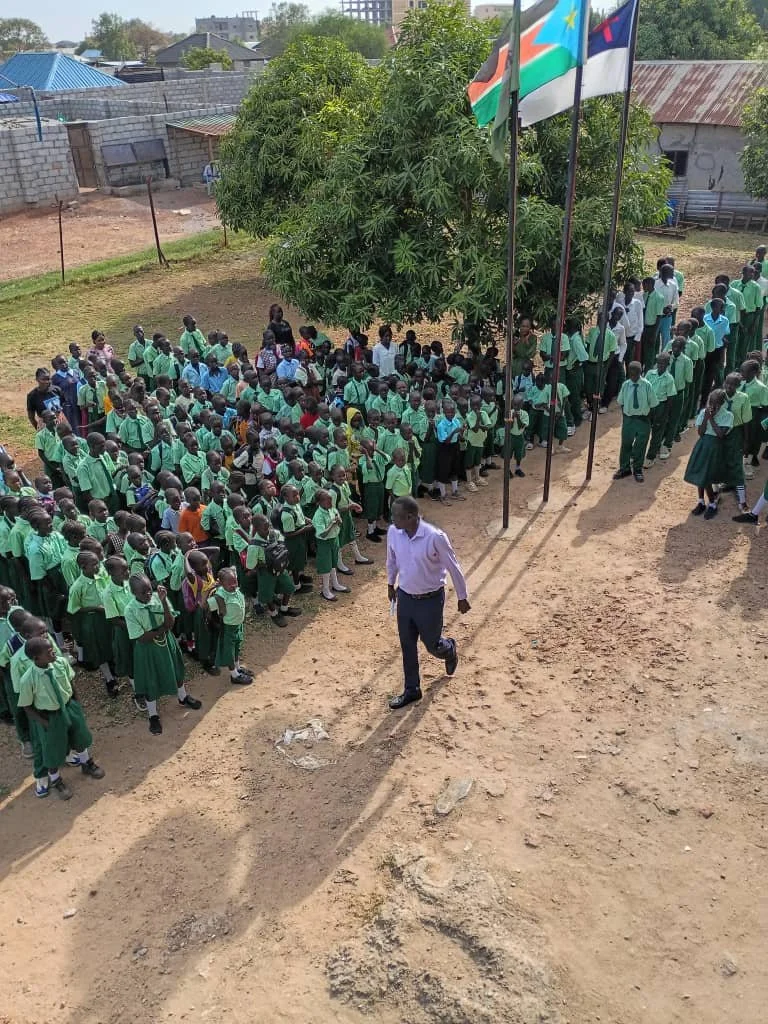 A group of school children in green uniforms standing in lines outdoors, with a man walking nearby. Four flags are flying on poles next to a large tree, with a classroom or building in the background.