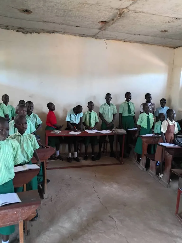 A classroom with a group of students standing behind desks, some with open notebooks, wearing green uniforms, in a basic room with a cracked concrete floor and unfinished ceiling.
