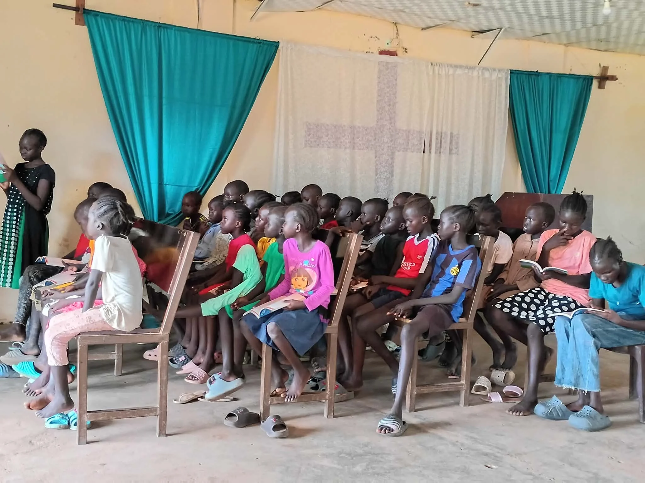 Children sitting on wooden chairs inside a classroom, facing forward, with a blue curtain and a white cloth backdrop at the front. Some children are reading books, and their shoes are removed and placed on the ground.