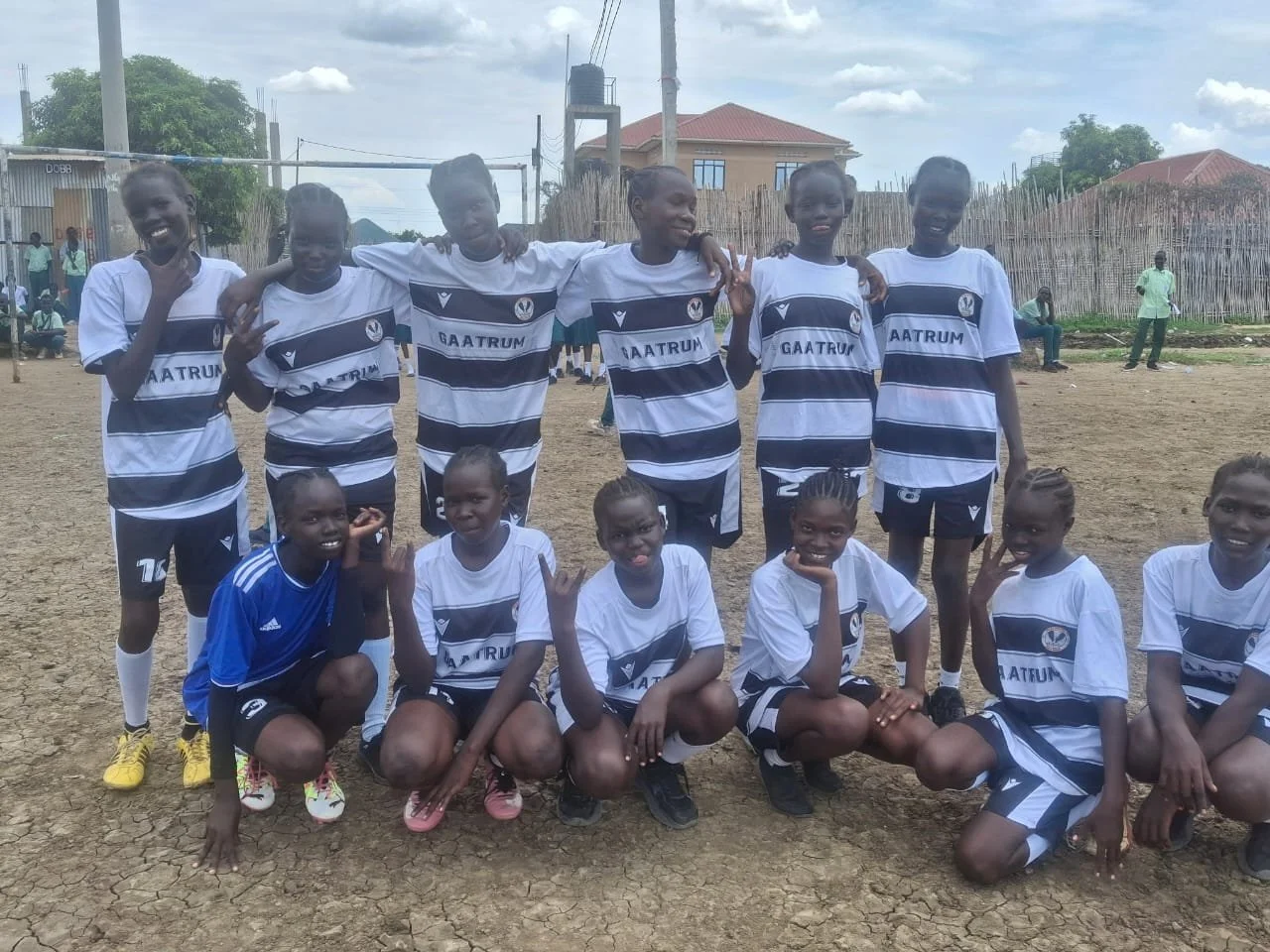 Group of young girls on a dirt football field, wearing soccer uniforms. Some are smiling and making peace signs, others are kneeling or standing with arms around each other. Background shows wooden fences, houses, and a cloudy sky.