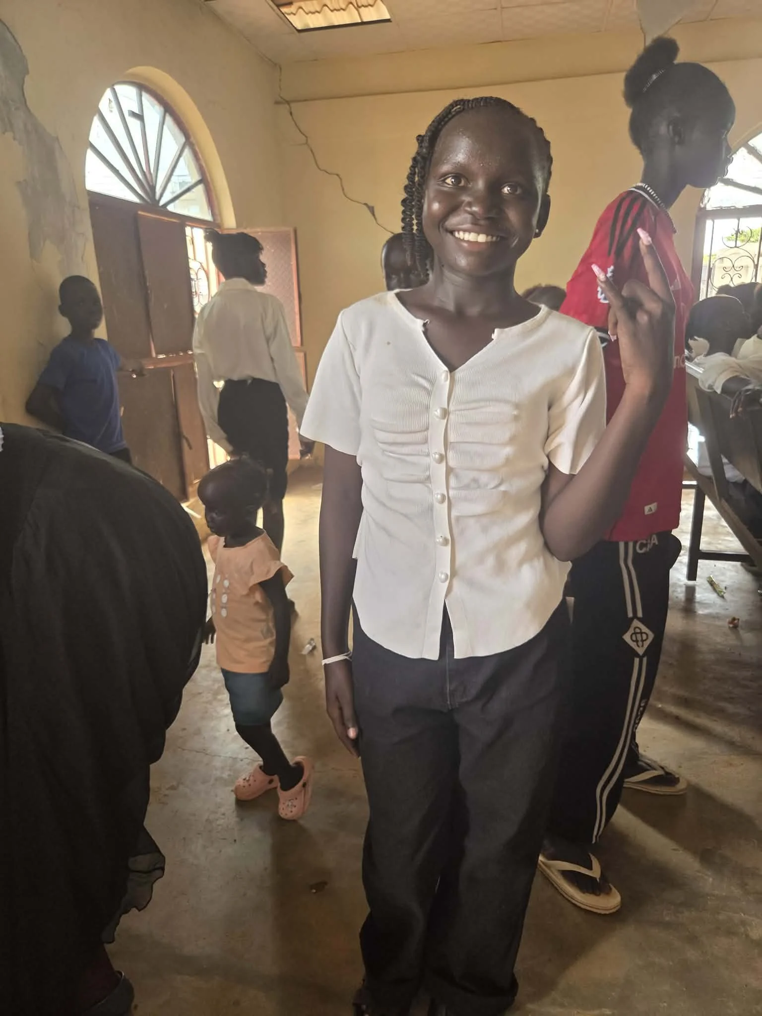 Smiling woman in white blouse standing indoors with other people in the background, some looking outside, in a room with cracked walls and a partially open window.