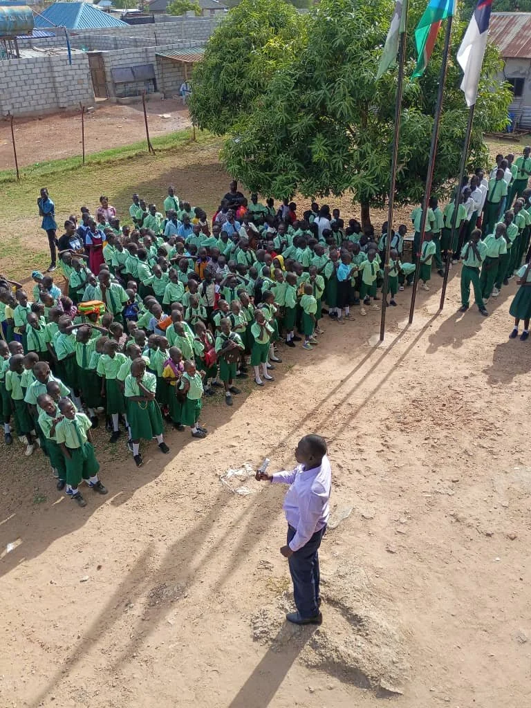 A man standing outside in front of a large group of school children, who are wearing green uniforms, gathered around a flagpole under a tree. The scene appears to be a school assembly or flag-raising ceremony.