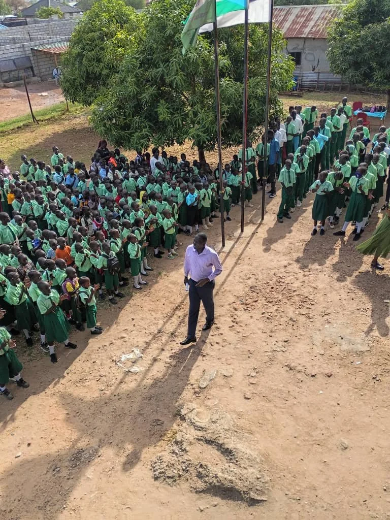 A large group of school children in green uniforms are standing in lines outdoors, with a man in a white shirt and dark pants in front of them. There are three flagpoles with flags and trees around. The setting appears to be a schoolyard in a rural o