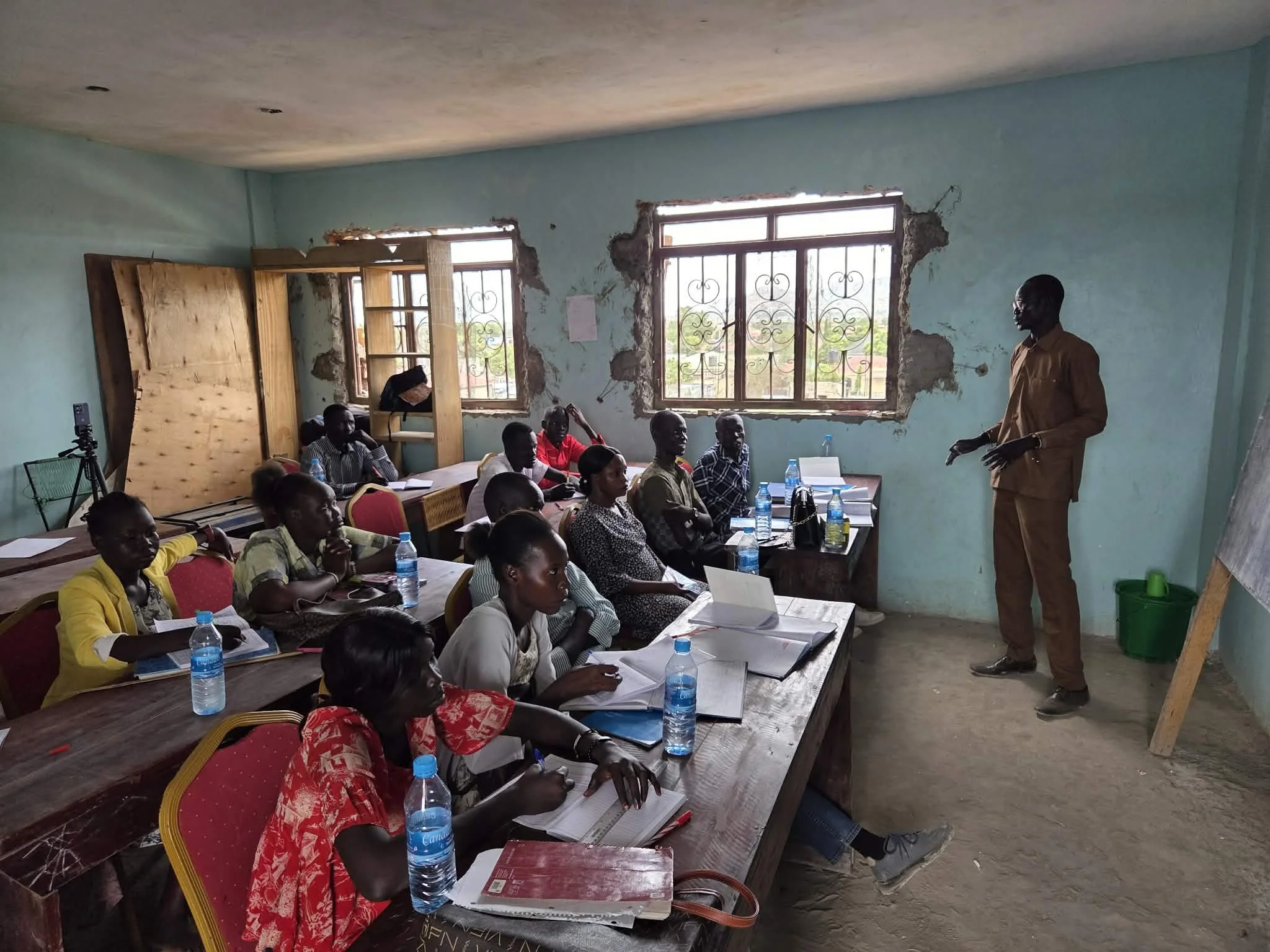 A classroom with students seated at desks, listening to a male teacher standing by the wall. The room has three windows with decorative iron bars, and the walls are partially damaged. Bottles of water and notebooks are on the desks.