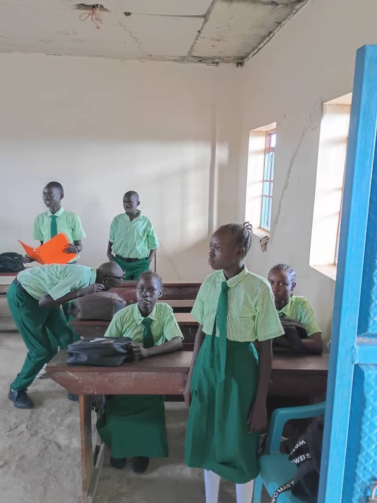 A classroom with five young students in uniform, four sitting at desks and one standing, with three other students in the background, in a worn room with a damaged ceiling and a window on the right.