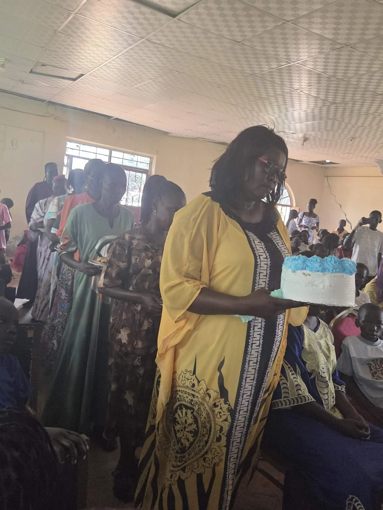 A woman in glasses wearing a yellow traditional dress holds a cake with blue cream in a room filled with people, some sitting and some standing, during a celebration or gathering with natural light coming from the window.