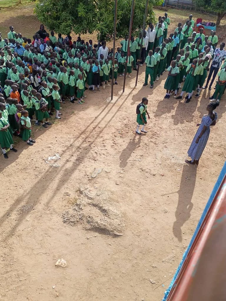 School children in green uniforms gathered outside in a courtyard, some standing in lines, with teachers supervising.