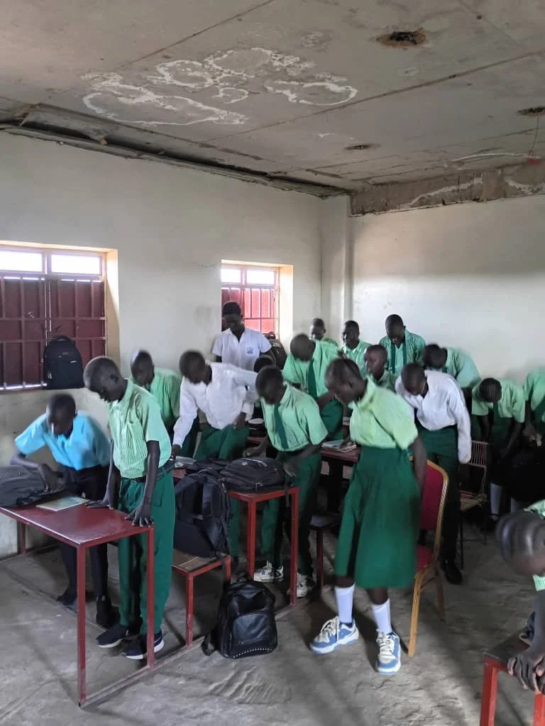 Students in green and white uniforms standing and bowing in a classroom, with backpacks on desks and the floor, and windows in the background.