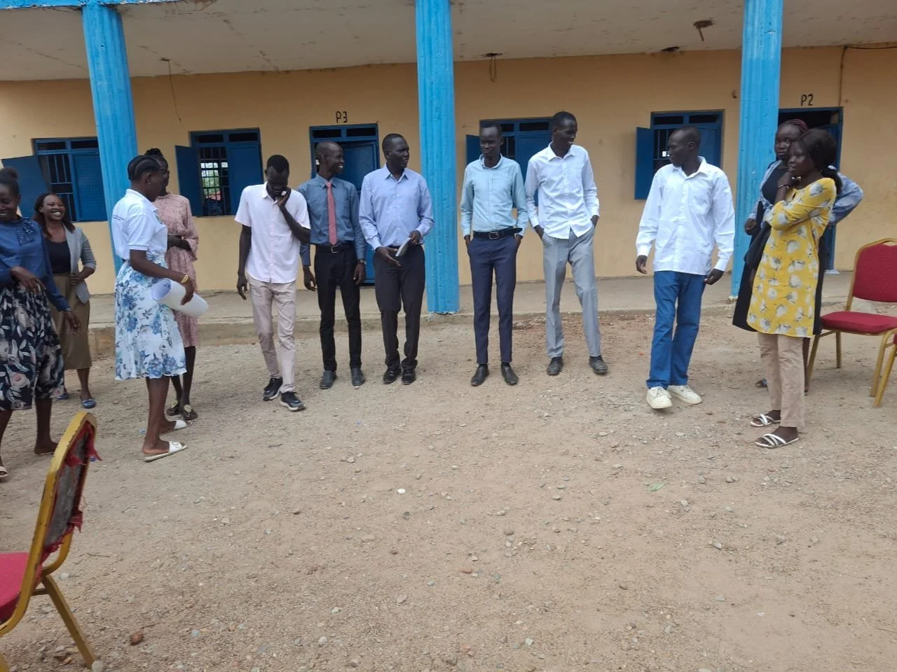 Group of people standing in a line outdoors near a building with blue window frames and pillars, some smiling and engaging in conversation.