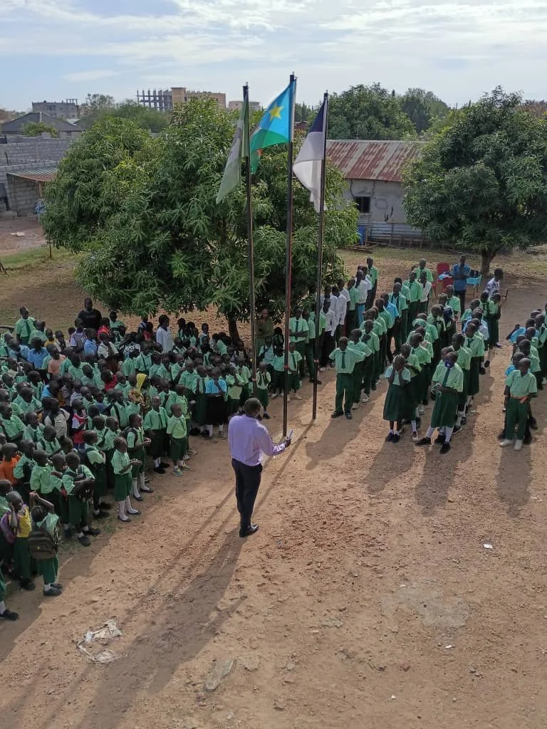 A school assembly with students standing in rows on a dirt ground around a flagpole with three flags, including a flag with a blue background and yellow star, under a large tree, in a rural area.