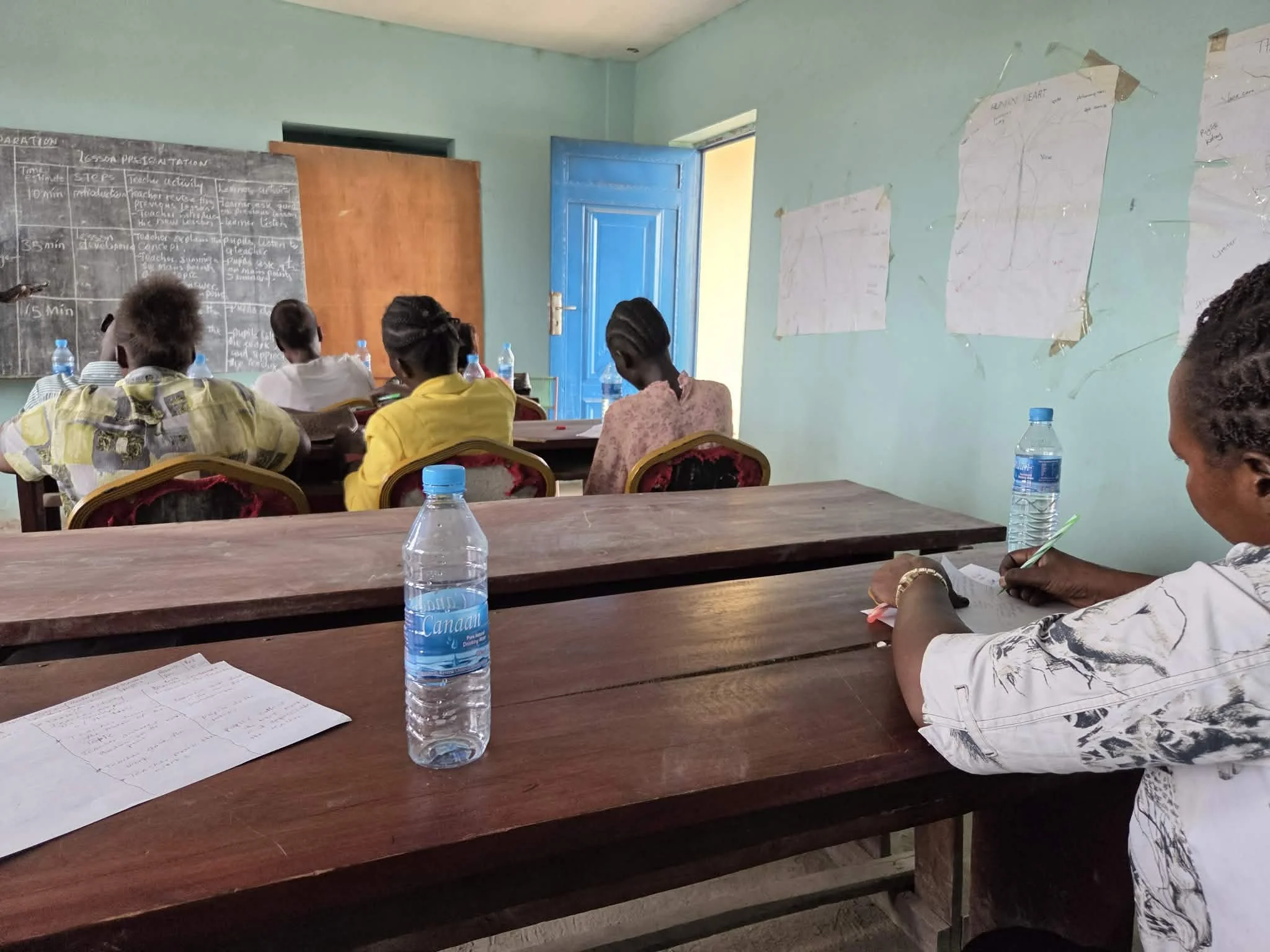 Classroom with students seated at desks, water bottles, notebooks, a blackboard with writing, and posters on the wall, with a blue door open to the outside.