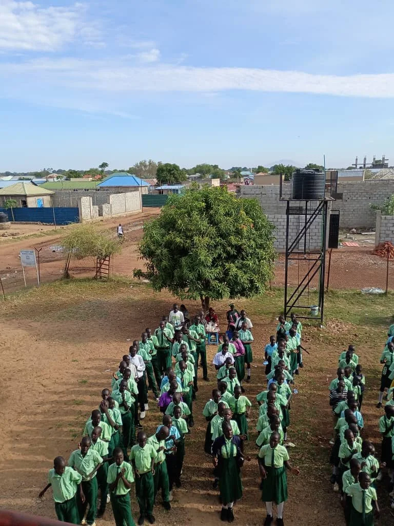 Children in green uniforms standing in orderly lines outside in a schoolyard, with a large tree, water tank, and buildings in the background.
