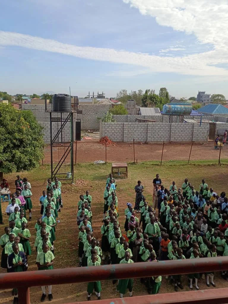 Students standing outside on a school yard, organized in rows, with some standing near desks and others in a group, during daytime under partly cloudy sky.