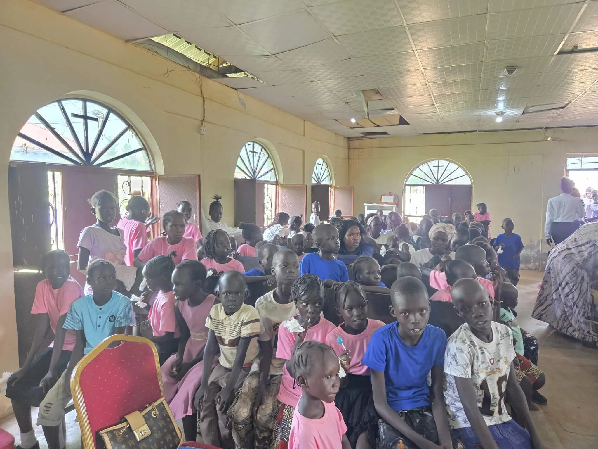 A large group of children gathered inside a building, some sitting and some standing, with a few adults also present. The room has arched windows and a decorated ceiling, and the children are wearing colorful clothing.