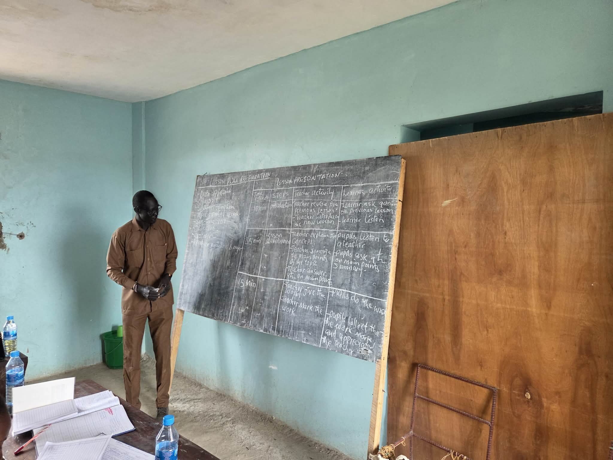 Man in brown shirt standing in classroom near chalkboard with lesson plan, several water bottles, and papers on desk, with green and wooden walls.