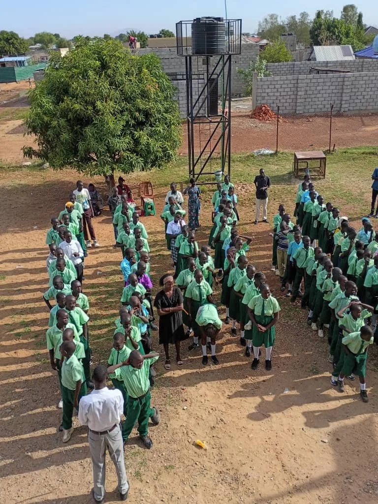 School children in green uniforms standing in lines on a dirt field, with teachers supervising, under a clear sky, with a water tank on a tall stand and a large tree nearby.