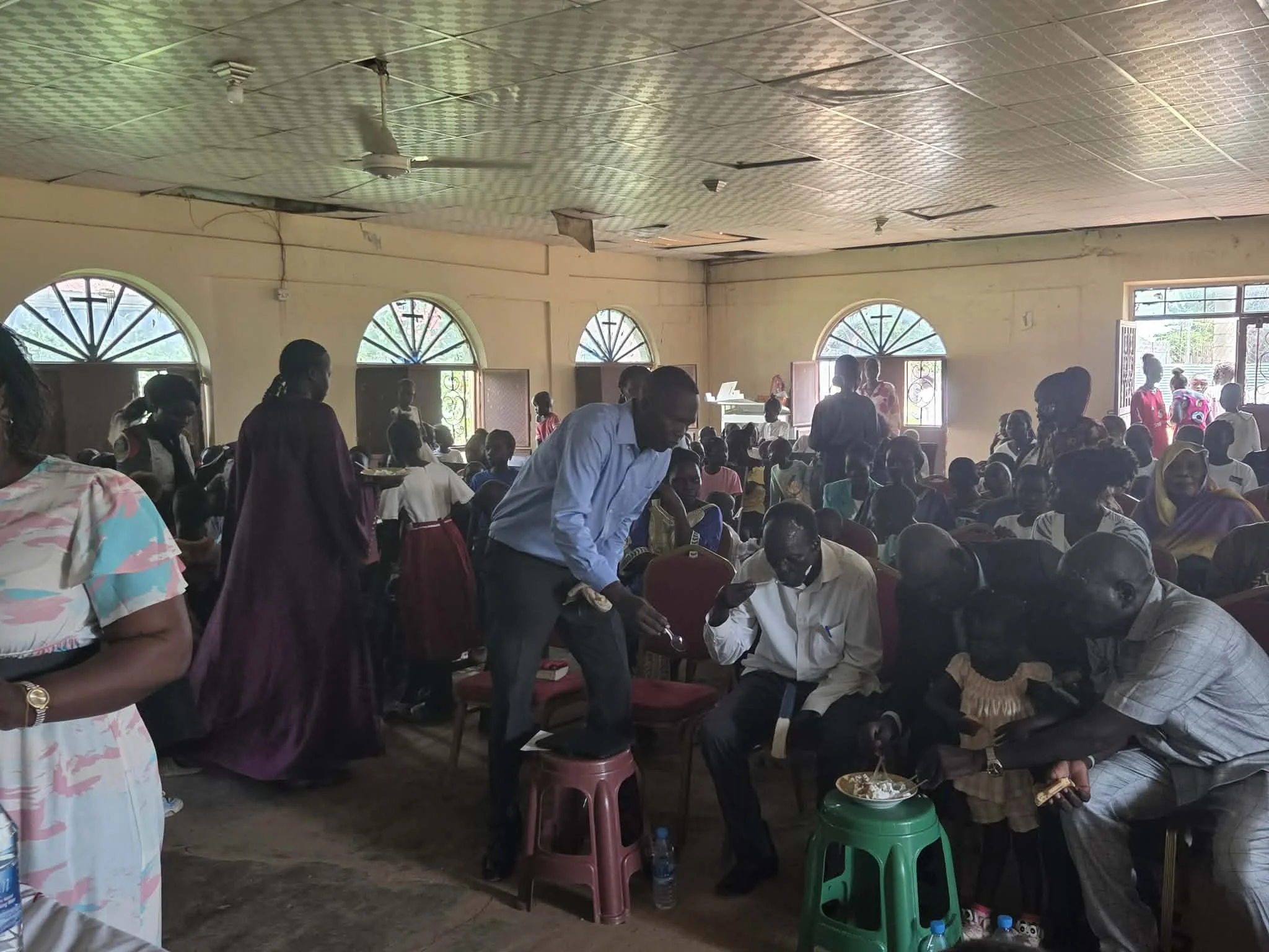 A crowded room filled with people, many seated and some standing, during an indoor gathering or event. The room has large arched windows, and some people are eating or holding plates, while others are engaging in conversations.
