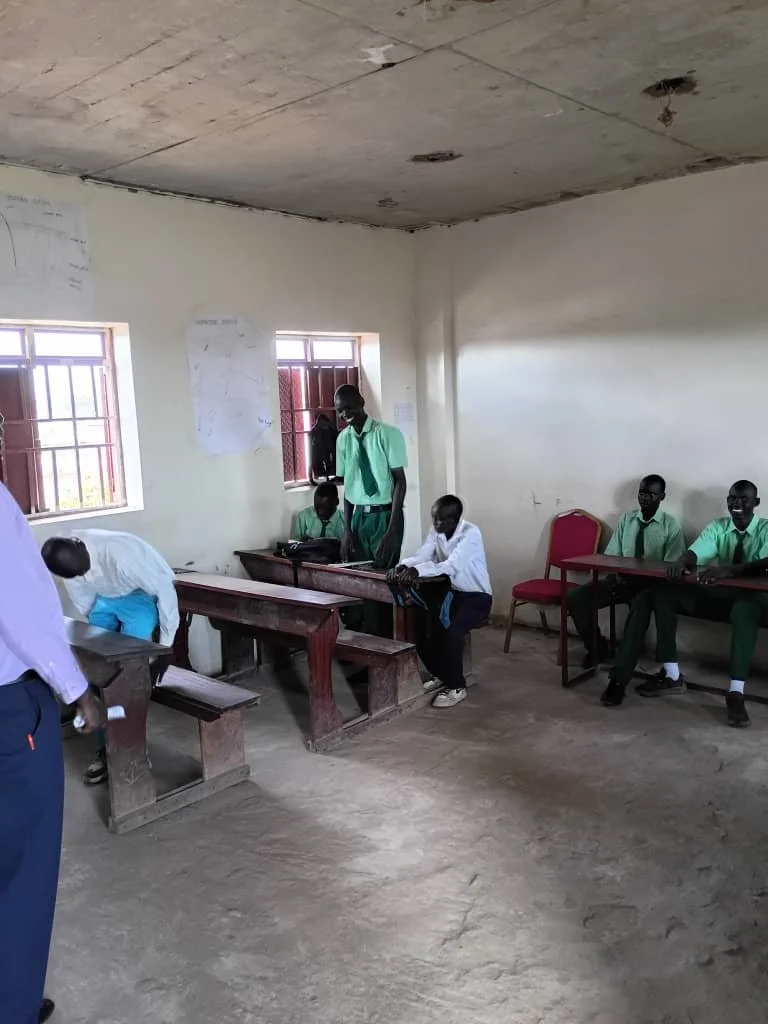 A classroom with six students, some sitting at desks and some standing, with a teacher in the foreground. The room has two windows, a plain wall, and a ceiling with visible damage.