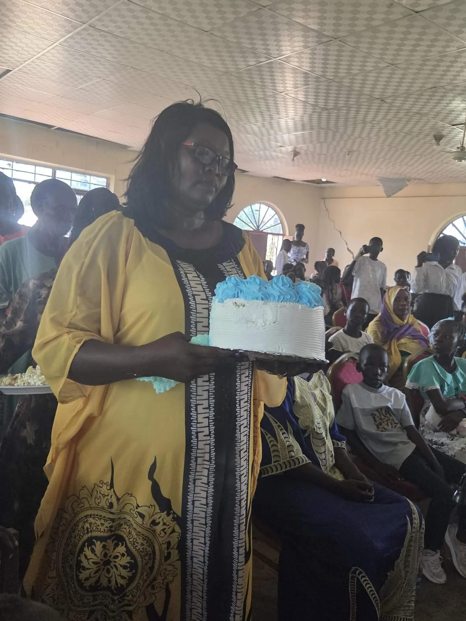 A woman in a yellow kimono holding a cake at a gathering inside a hall with many seated and standing people.