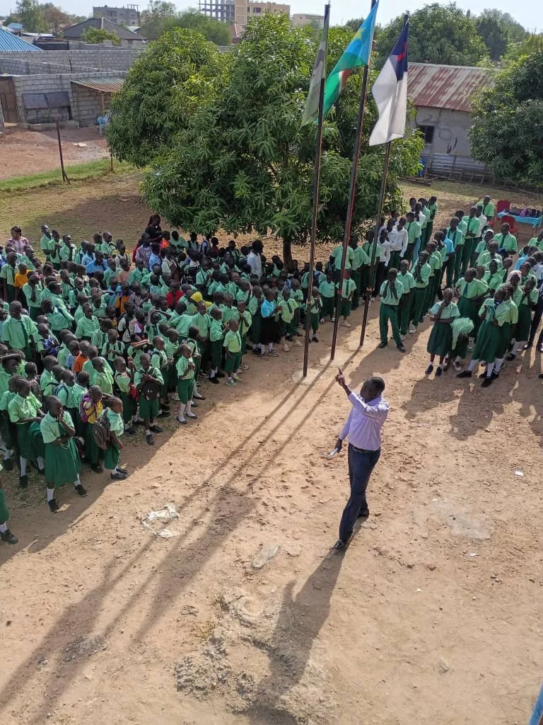 A large group of students in green school uniforms gathered outdoors, standing in lines under a tree, with a teacher or supervisor speaking to them. Flags are flying on poles near the tree on a sunny day.
