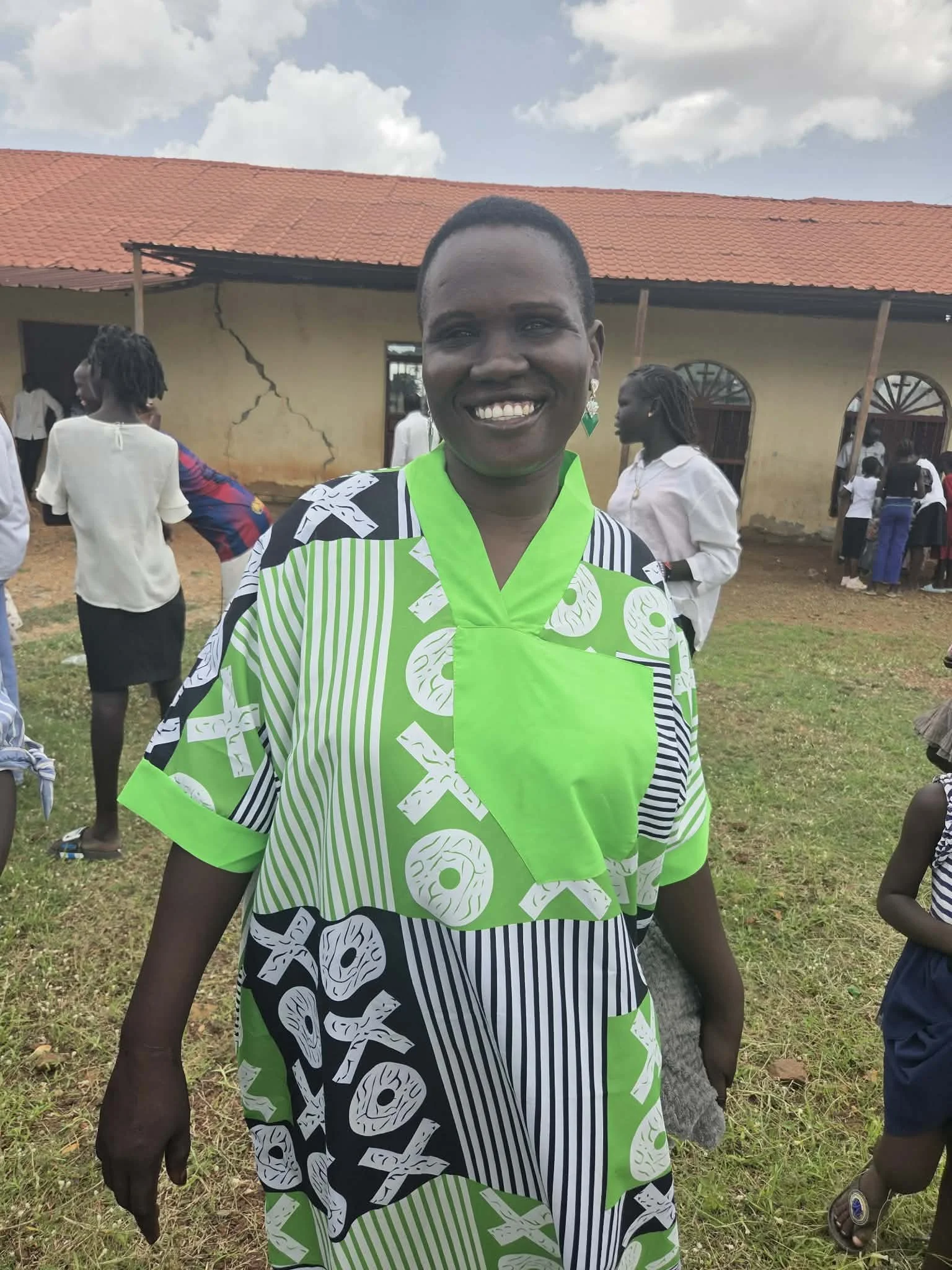 A woman smiling at an outdoor gathering, wearing a green, black, and white patterned shirt and earrings, with a boy in striped clothing nearby, in front of a building with a cracked wall and a red tile roof.