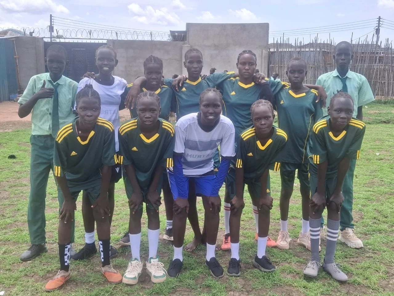 A group of young soccer players and two boys in casual clothing, posing on a grassy field with a concrete wall, fencing, and buildings in the background.