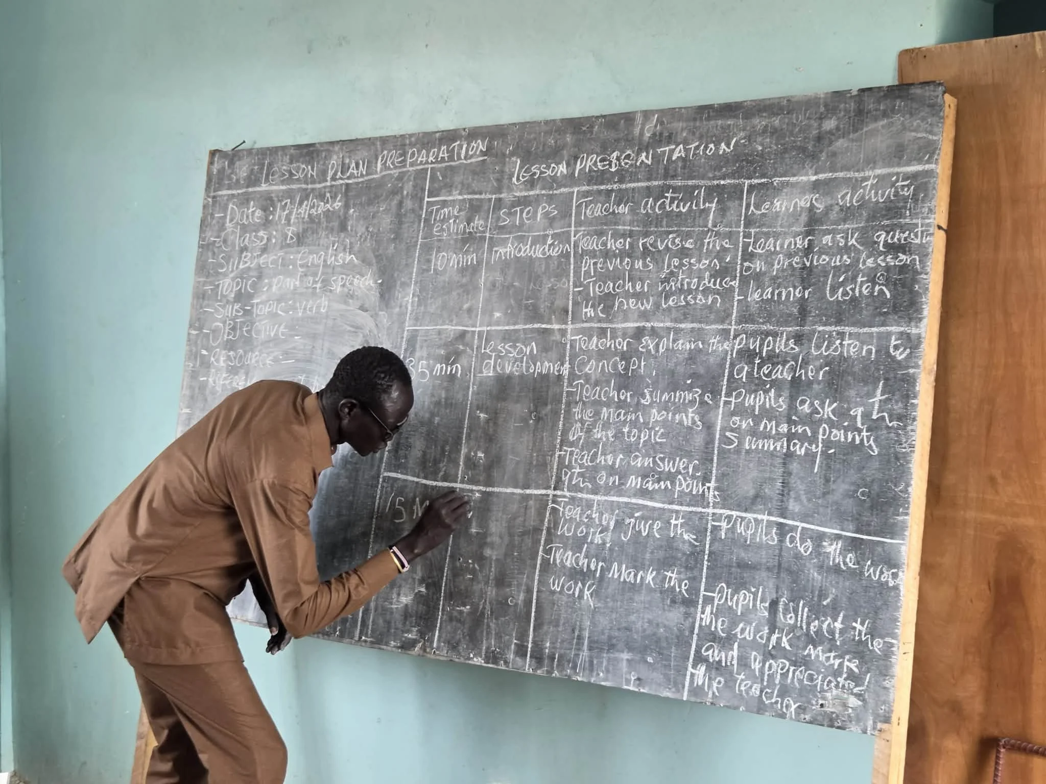 A man wearing glasses and a brown shirt writing on a blackboard in a classroom.