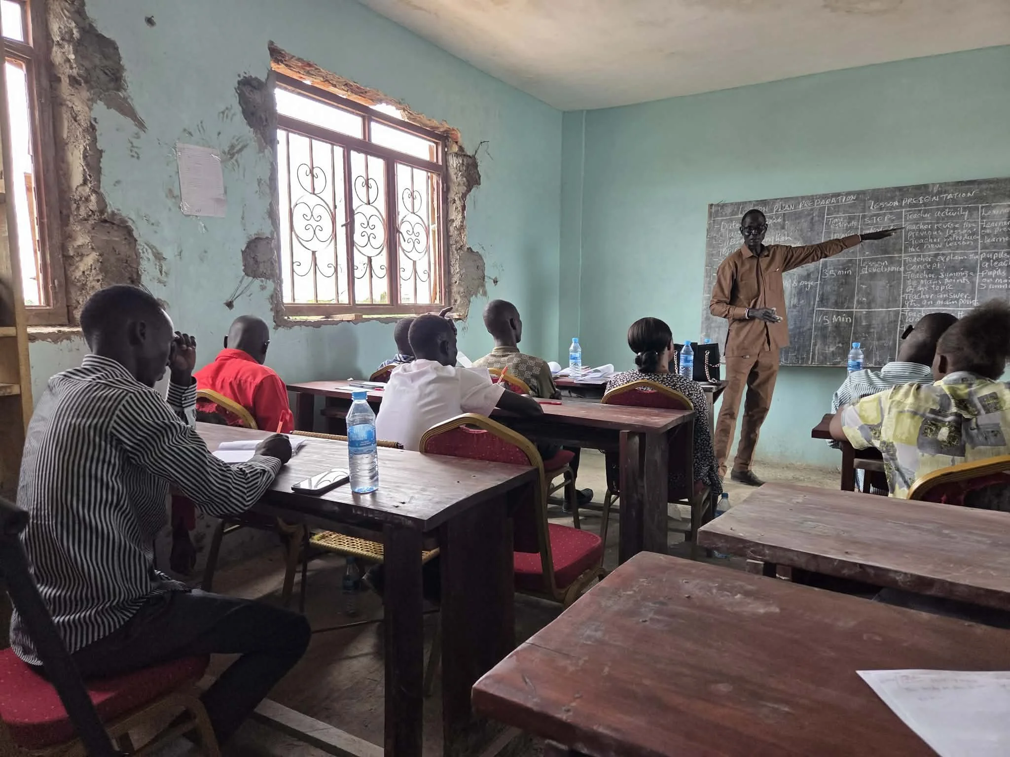 A classroom with students seated at desks, listening to a male teacher pointing at the blackboard. The room has two windows with decorative grillwork, and the walls are painted light green.