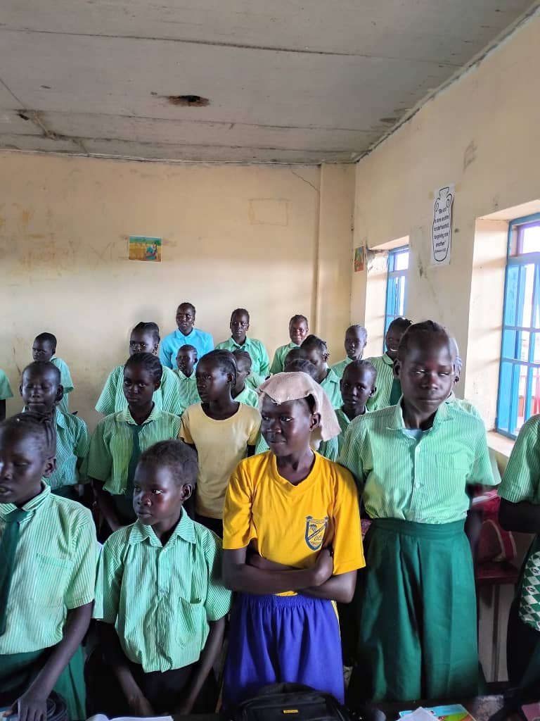 Group of school children standing in classroom with yellow, green, and blue uniforms.
