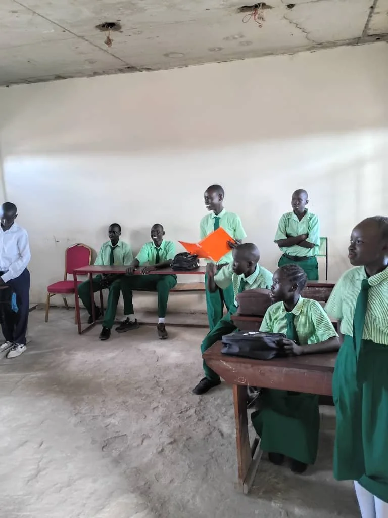 A group of schoolchildren in uniform are gathered in a classroom, some standing and some sitting at desks, with a plain wall and unfinished ceiling in the background.