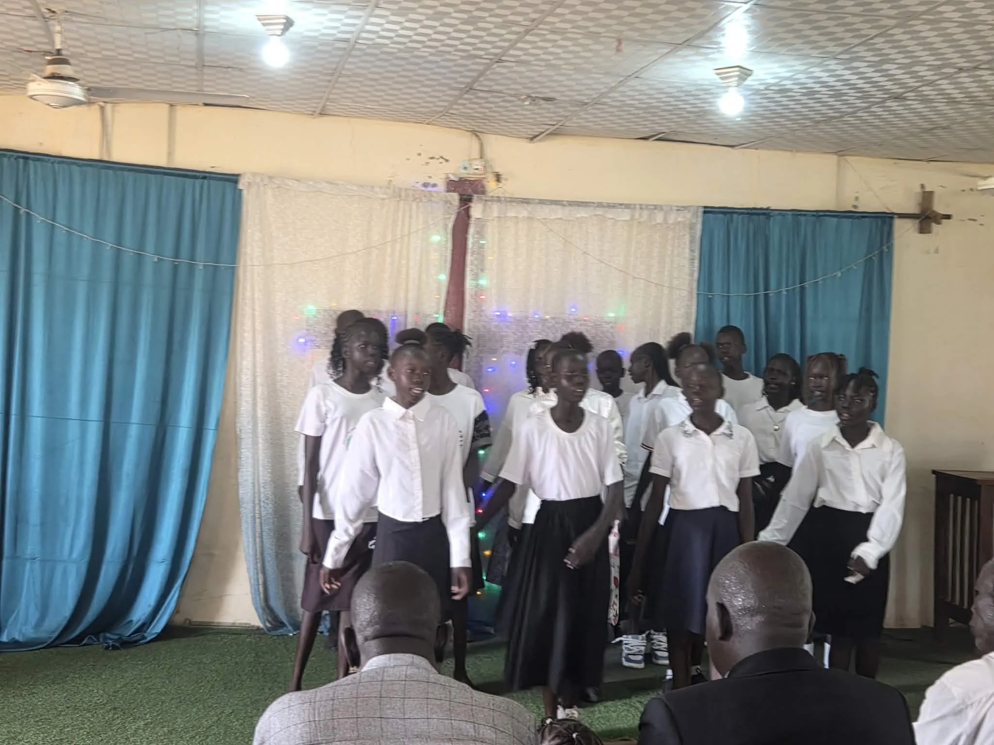 A group of people, mostly women, standing together in white shirts and dark skirts, are gathered indoors during a event. Behind them, there are blue curtains and colorful string lights. Some individuals are smiling, and there are people seated in fro