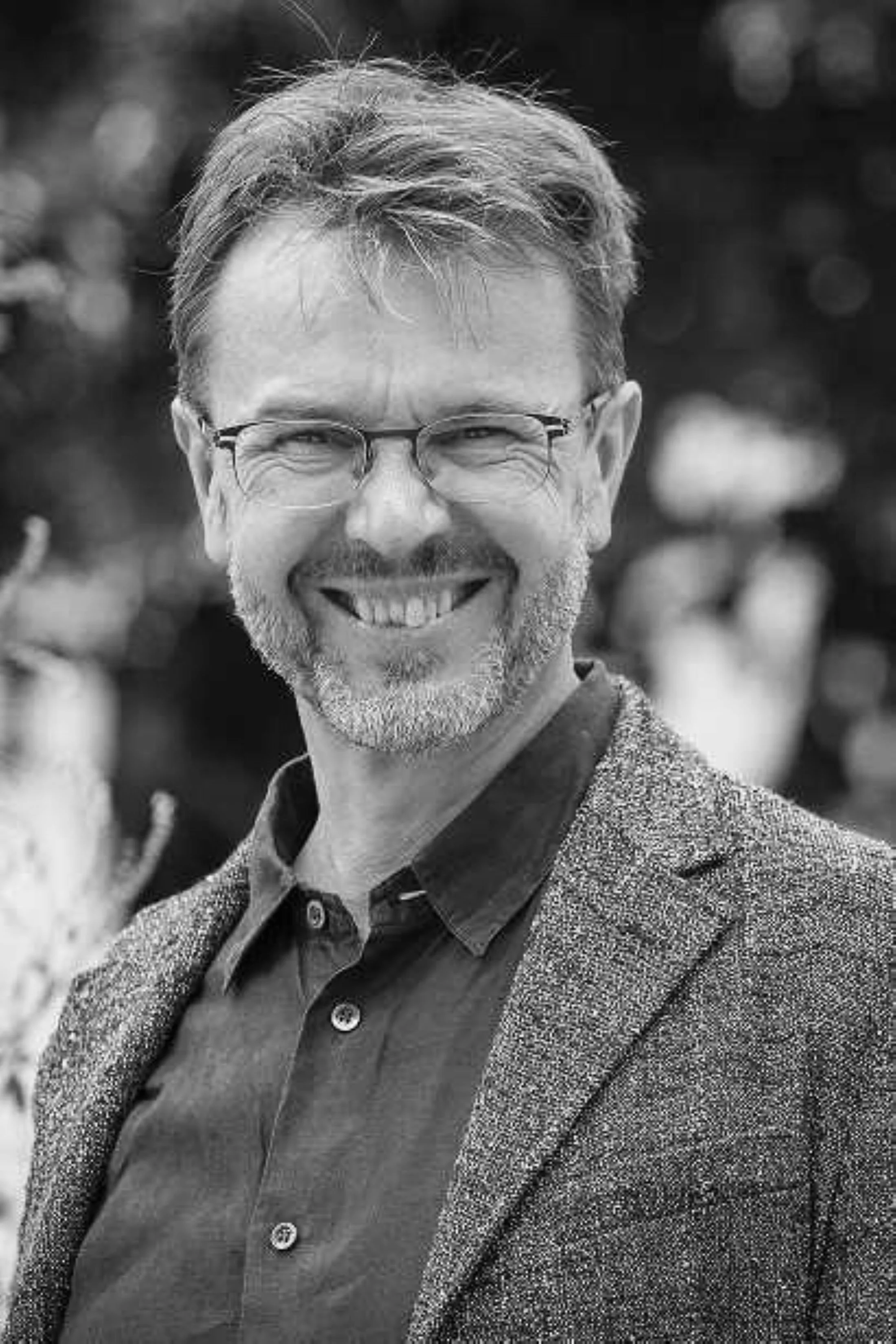 Schwarz-Weiß-Foto eines lächelnden Mannes mit Brille, grauem Haar und Bart, trägt einen Blazer und ein Hemd, im Hintergrund unscharfer Garten oder parkähnliche Umgebung.
